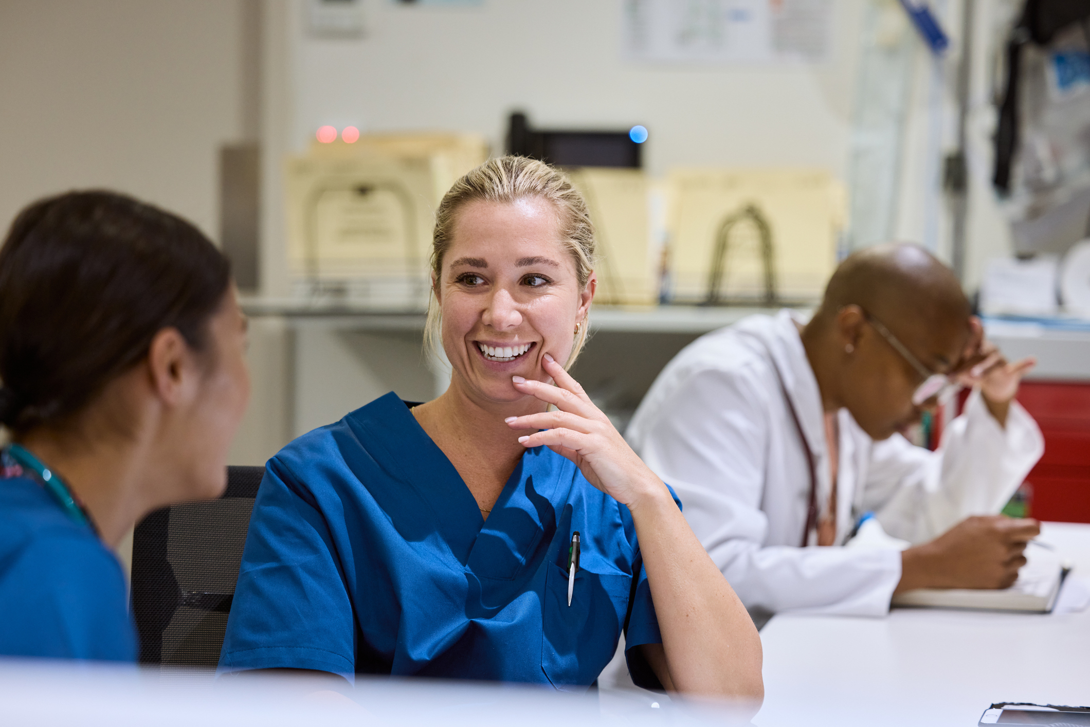 Smiling nurse chatting with colleague in hospital. Medical staff enjoying friendly conversation during break. Female healthcare workers are in blue scrubs.