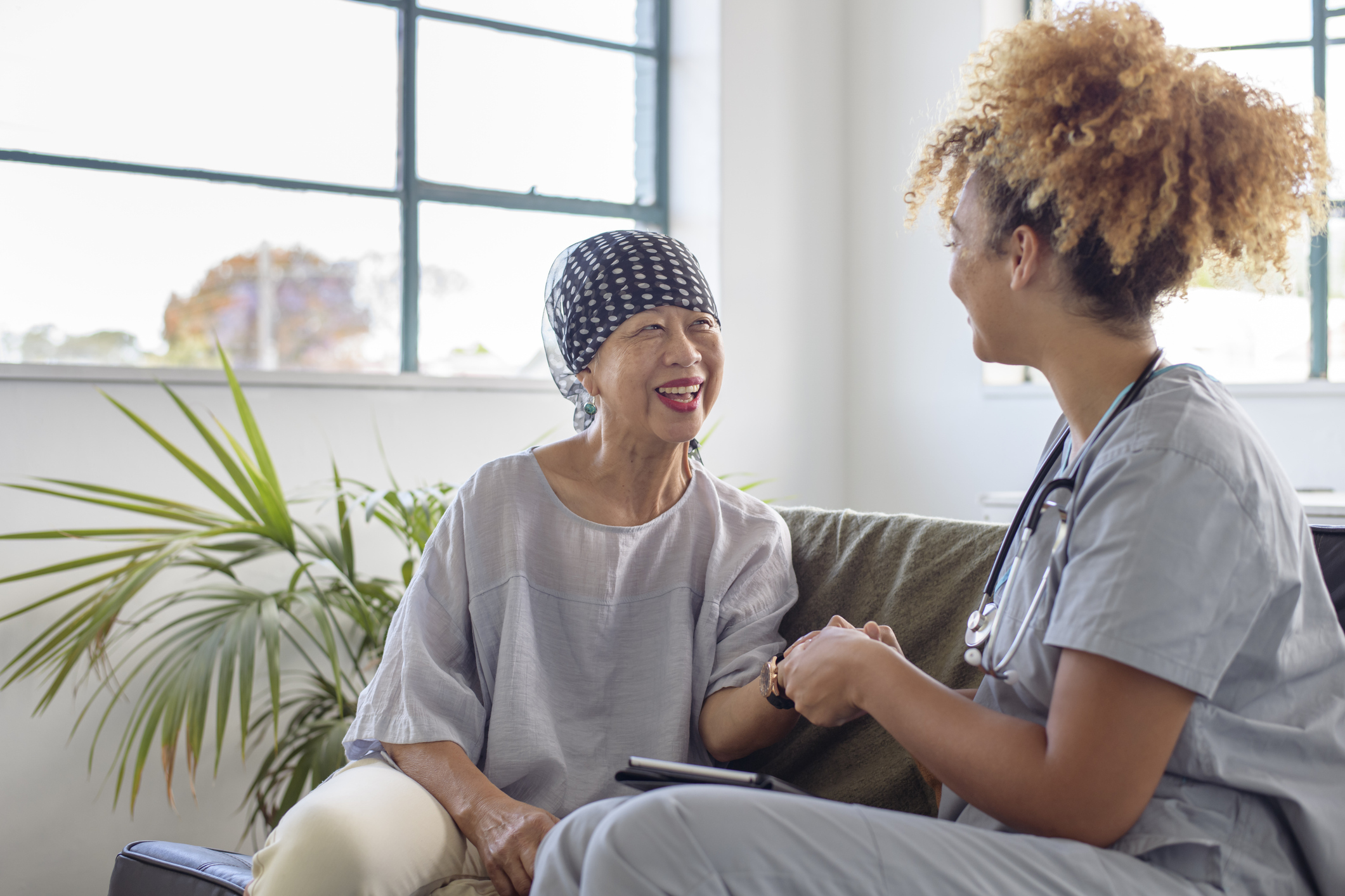 A senior woman of Asian descent smiles while having a casual conversation with her mixed race nurse who is providing in person care. The two women are siting side by side on a sofa in the patient's home.